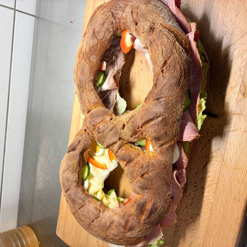 Ein Sandwich in Form der Zahl Acht, gefüllt mit Schinken, Käse und Gemüse, auf einem Holzbrett platziert.