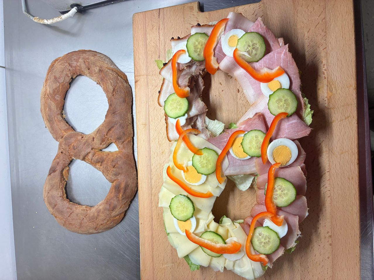 Ein kreisförmiges Sandwich auf einem Holzbrett mit Schinken, Eiern, Gurken und roten Paprika, geformt wie die Zahl 8. Daneben ein runder Brezel.