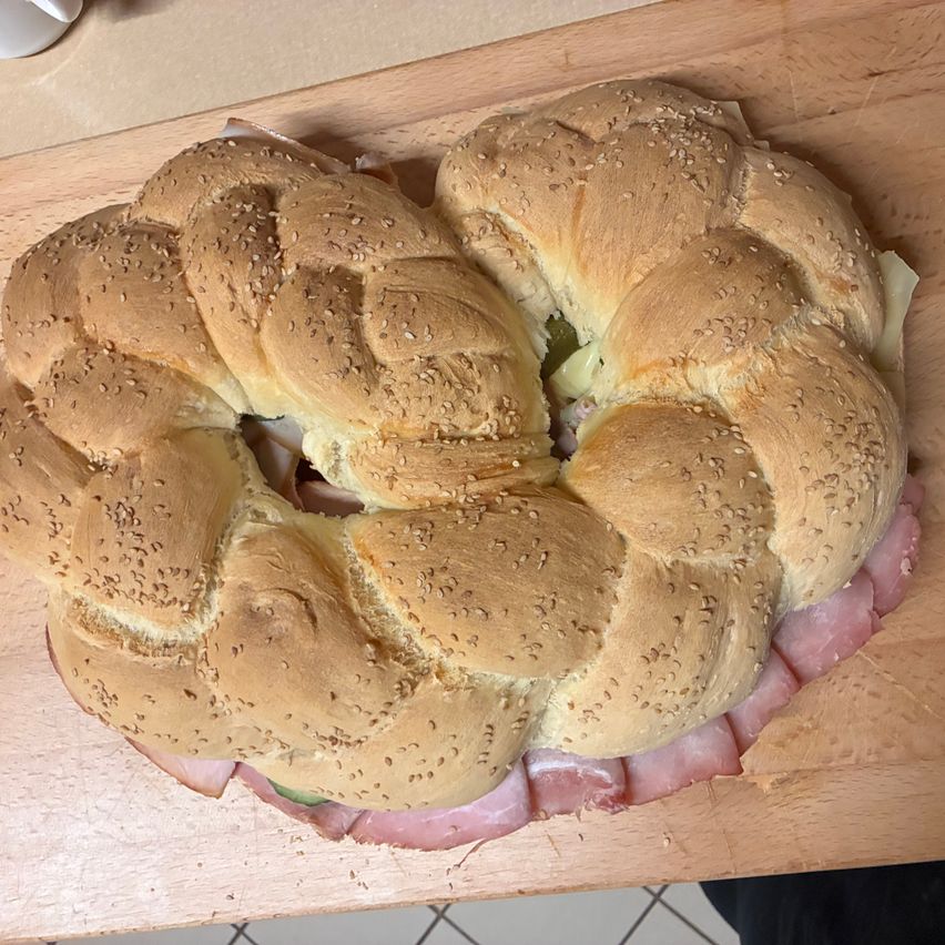 Ein kreisförmiges Sandwich mit Sesamkörnern auf einem Holzbrett, mit Schichten aus Schinken, Käse und Essiggurken. Das Sandwich ist in der Mitte aufgeschnitten und zeigt seine Füllung.