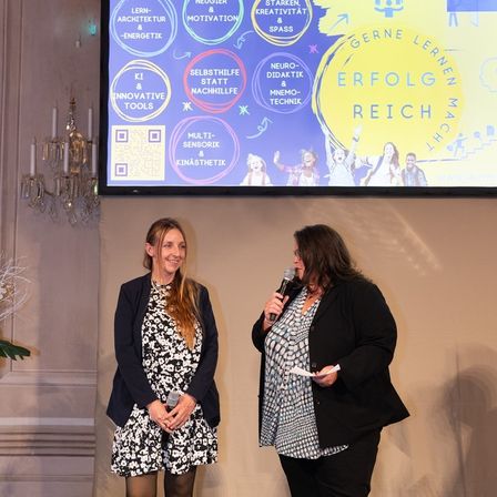 Zwei Frauen auf der Bühne, eine spricht in ein Mikrofon, die andere hält ein Papier, dahinter zeigt eine große Leinwand Bildungsinhalte an.