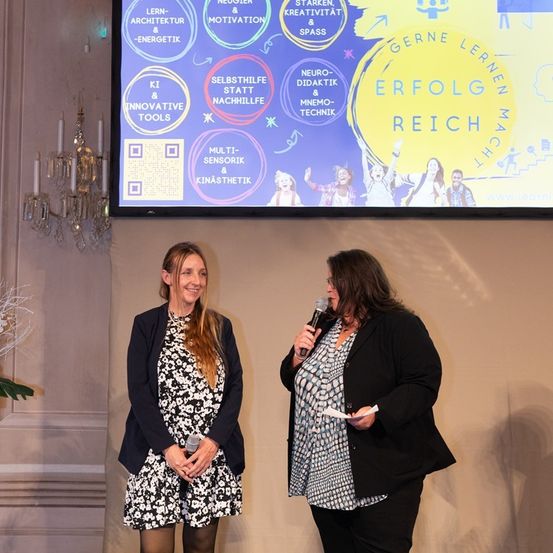 Zwei Frauen auf der Bühne, eine spricht in ein Mikrofon, die andere hält ein Papier, dahinter zeigt eine große Leinwand Bildungsinhalte an.