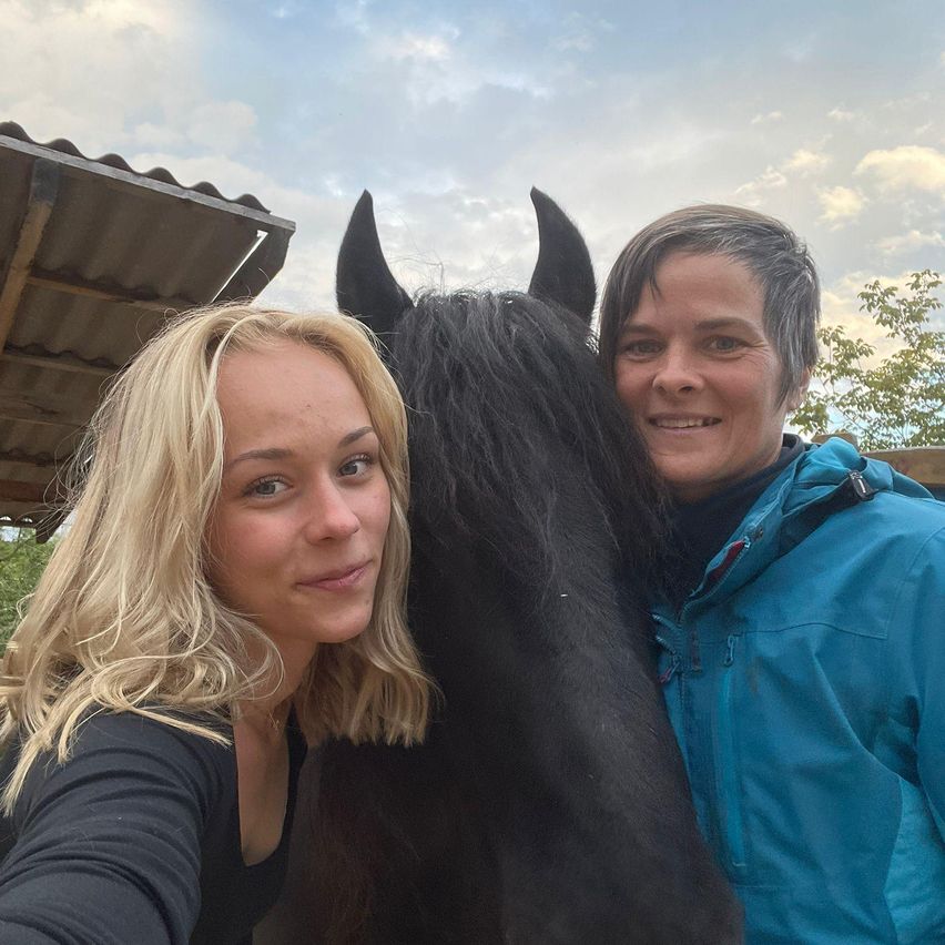 Zwei Frauen stehen neben einem schwarzen Pferd, eine in einem schwarzen Shirt und die andere in einer blauen Jacke. Sie lächeln und posieren für ein Foto.