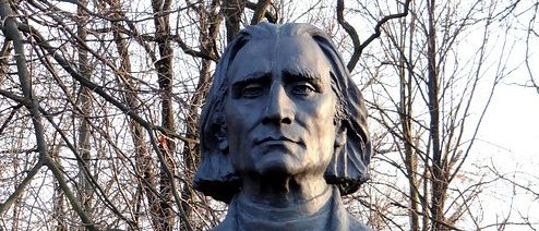 Eine Statue eines Mannes steht in einer Außenumgebung, wahrscheinlich in einem Park. Er ist in einem formellen Mantel gekleidet und hat lange, wellige Haare. Hinter ihm sind kahle Bäume, und der Himmel ist klar.