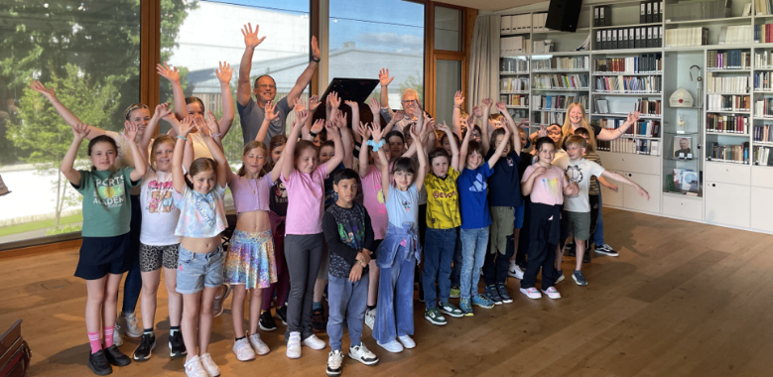 Eine Gruppe von Kindern und Erwachsenen steht in einem Raum mit erhobenen Händen und lächelt für ein Foto. Dahinter hält eine Frau einen Laptop. Der Raum hat große Fenster und Bücherregale.