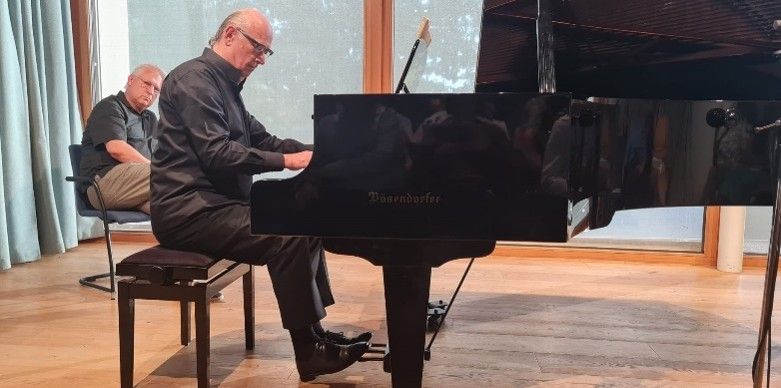 Ein älterer Mann spielt ein Flügel in einem Raum mit Holzböden und Glasfenstern. Ein anderer Mann sitzt in einem Stuhl hinter ihm und beobachtet.