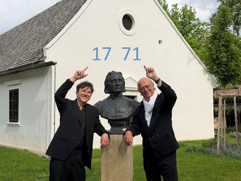 Zwei Männer in Anzügen posieren neben einer Büste eines Mannes vor einem weißen Gebäude mit den Zahlen 17 71 darüber.