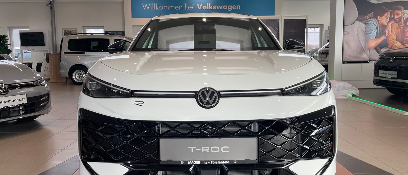 Ein weißer Volkswagen T-Roc wird in einer Ausstellungshalle präsentiert, mit einem blauen Schild, das 'Willkommen bei Volkswagen' steht.