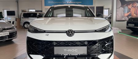 Ein weißer Volkswagen T-Roc steht in einer Autoausstellung, an einer blauen Tafel mit der Aufschrift 'Willkommen bei Volkswagen.'