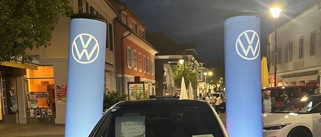 Ein silberner Volkswagen ID.3 Neo Elektroauto steht nachts auf der Straße mit zwei blauen Volkswagen-Bannern im Hintergrund.