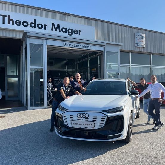 Mehrere Personen stehen neben einem weißen Audi-Auto vor einem Gebäude mit dem Schild 'Odor Mager'.