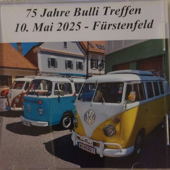 Ein Plakat zeigt drei alte VW-Bullis, zwei blaue und einen gelben, vor Gebäuden geparkt, mit einer Nachricht über ein 75-jähriges Treffen in Furstenfeld am 10. Mai 2025.