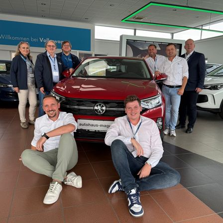 Eine Gruppe von Menschen posiert mit einem roten Volkswagen-Auto in einer Ausstellung. Sie tragen alle Ausweise und lächeln. Hinter ihnen stehen andere Autos und ein blaues Banner, das 'Willkommen bei Volkswagen' steht.