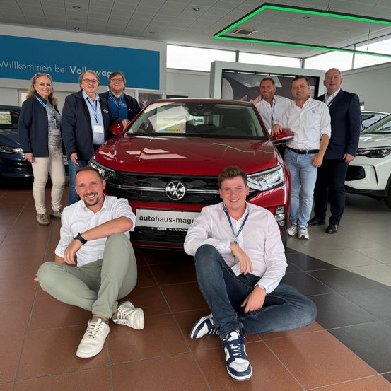 Eine Gruppe von Menschen posiert mit einem roten Volkswagen-Auto in einer Ausstellung. Sie tragen alle Ausweise und lächeln. Hinter ihnen stehen andere Autos und ein blaues Banner, das 'Willkommen bei Volkswagen' steht.