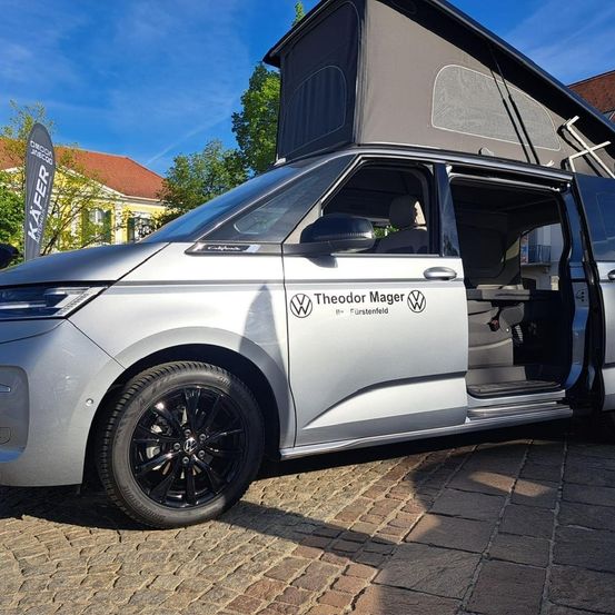 Ein silberner VW-Camperbus mit geöffneten Türen, vor einem Gebäude geparkt. Der Van hat ein graues Dach mit einer Markise und einem VW-Logo.