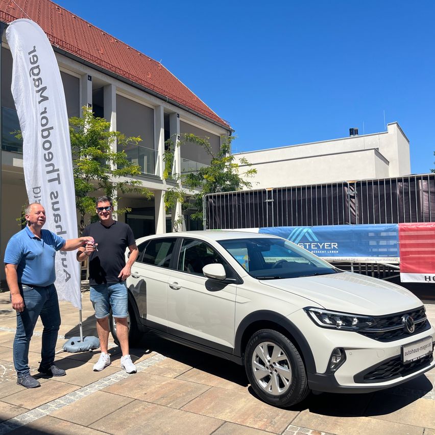 Zwei Männer stehen vor einem weißen Volkswagen Tiguan bei sonnigem Wetter. Ein Banner mit weißem Text steht hinter ihnen. Im Hintergrund sind Gebäude mit Glasfenstern zu sehen.