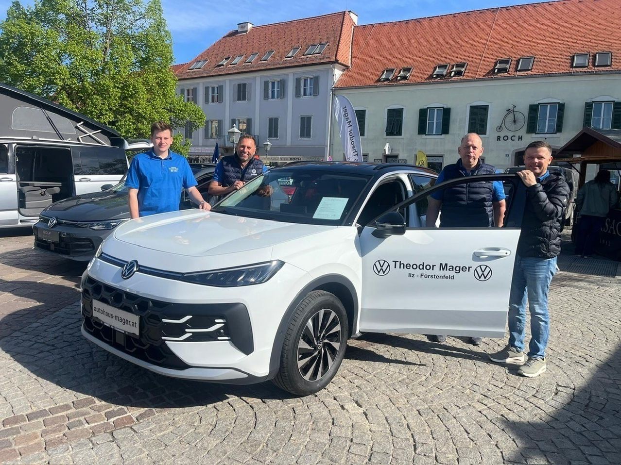 Vier Männer stehen vor einem weißen Volkswagen SUV. Einer hält die Tür offen, und ein Nummernschild liest 'autohaus-mager.at'. Dahinter ist ein Gebäude mit Fahrradschild sichtbar.