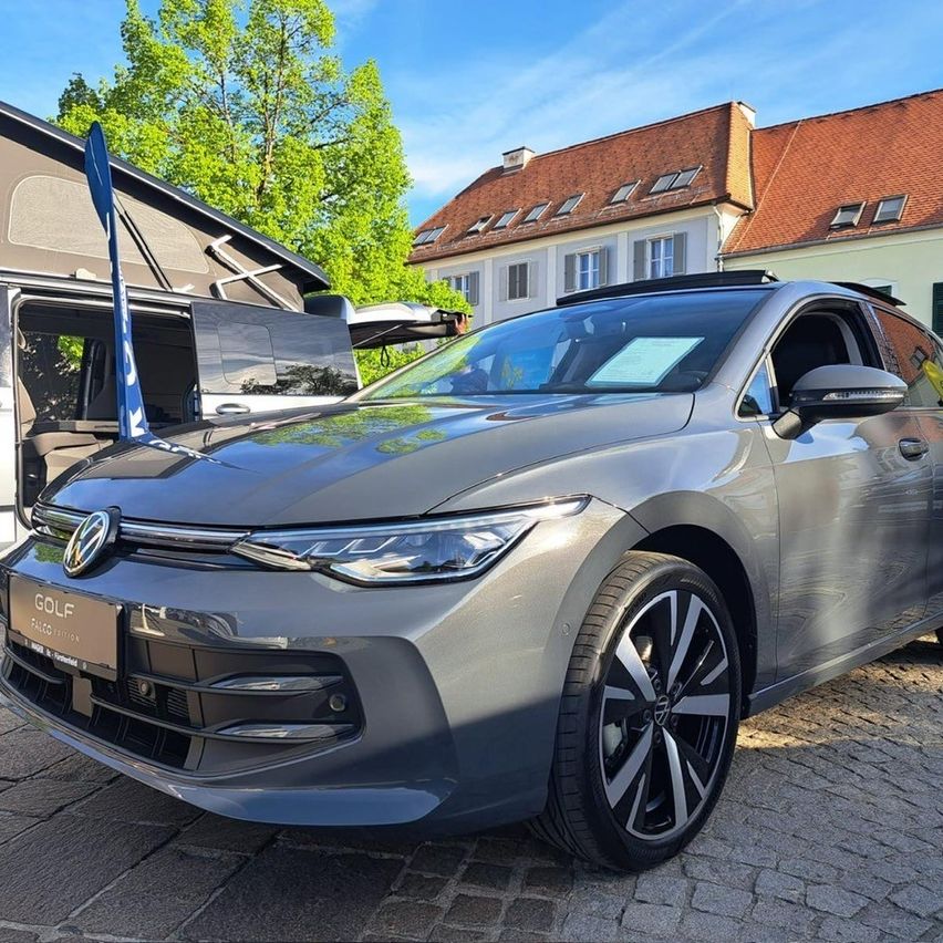 Ein Volkswagen Golf steht auf einer gepflasterten Straße vor einem Haus mit rotem Dach. Links ist ein Zelt zu sehen.