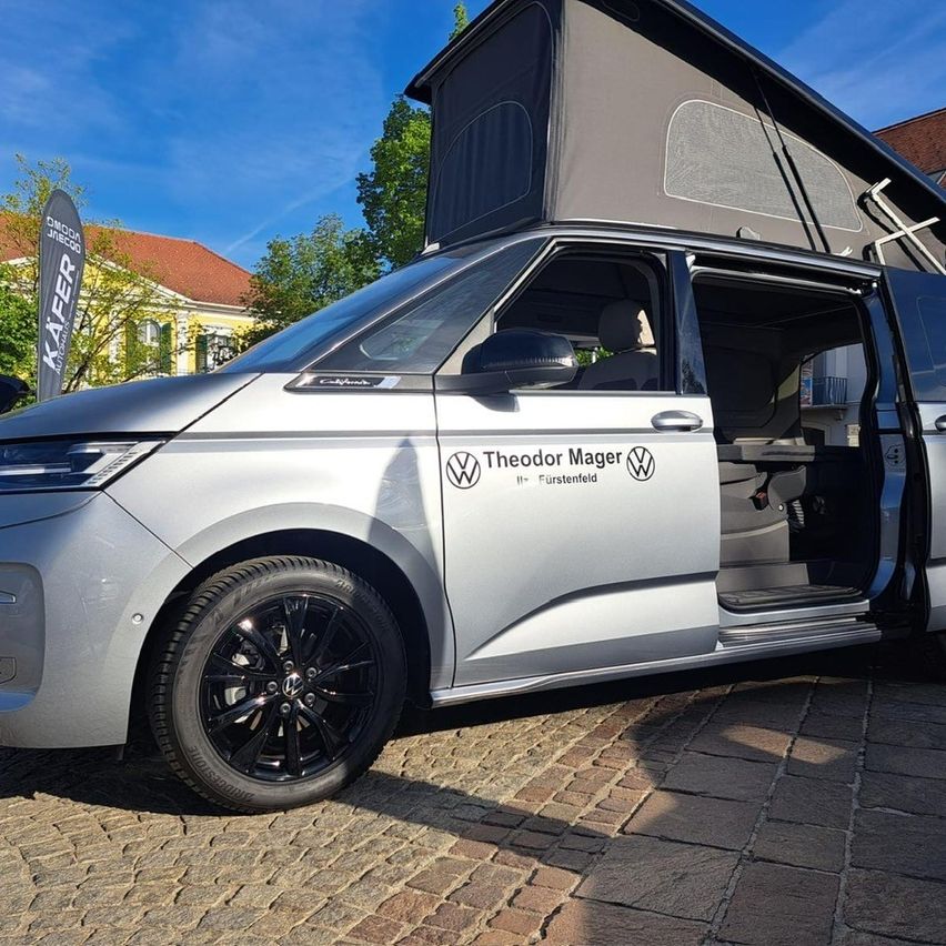 Ein silberner Wohnmobil mit erhöhtem Dach ist auf einem Ziegelweg geparkt und zeigt seine offenen Türen und das Theodor Mager Logo.