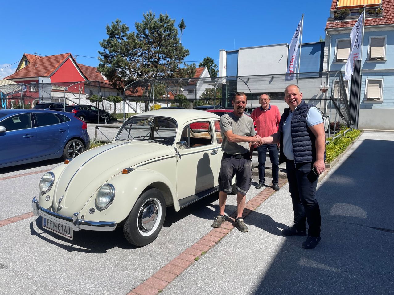 Three men are standing in front of a white vintage car with the license plate F461AU, shaking hands. Behind them, a building with a flag is visible.