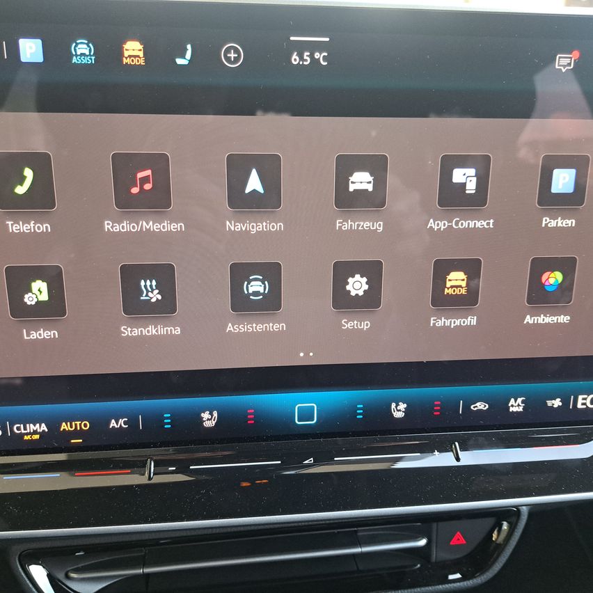Das Infotainment-Display eines Autos zeigt verschiedene Symbole für Funktionen wie Telefon, Radio/Medien, Navigation, Fahrzeug, App-Connect und Parken. Unten sind Symbole für Laden, Klima, Assistenz, Setup, Fahrprofil und Ambiente zu sehen.