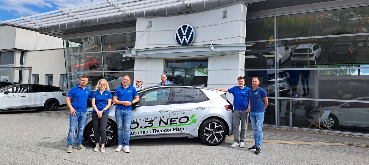 Fünf Personen stehen vor einem weißen Volkswagen-Auto mit dem Text D.3 NEO und einem grünen Blattsymbol an der Seite. Sie tragen alle blaue Hemden und lächeln. Dahinter befindet sich ein Gebäude mit einem Volkswagen-Logo oben.