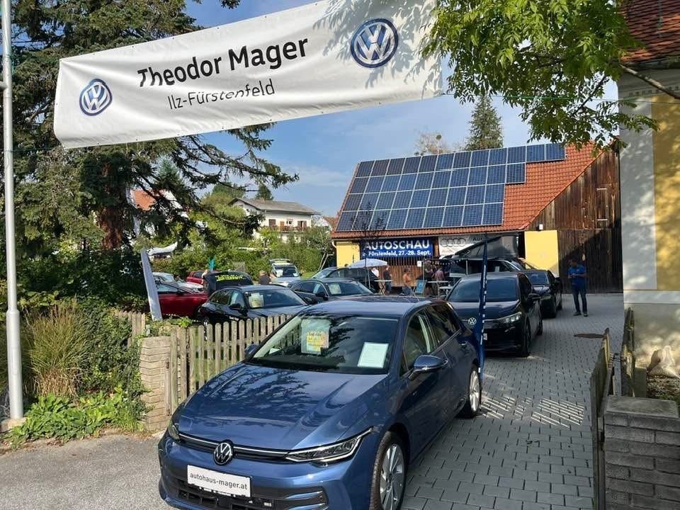 A blue Volkswagen car parked in front of a dealership with solar panels on the roof and a banner reading Theodor Mager Illz-Furstenfeld.
