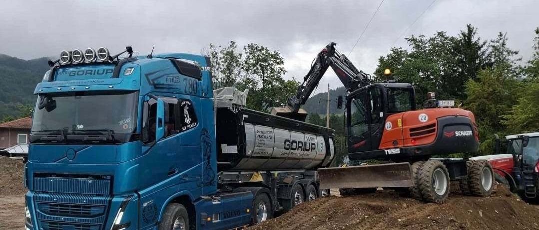Ein blauer Lkw mit dem Gorup-Logo ist auf einer Schotterstraße geparkt. Ein schwarzer und orangefarbener Bagger schiebt Erde von der Straße. Im Hintergrund befindet sich ein weiteres Fahrzeug.