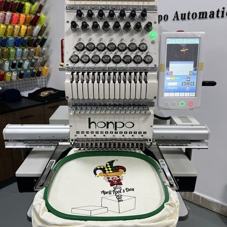 Eine Honpo-Stickmaschine stickt ein Design eines Narren auf einem Stoff. Die Maschine ist von verschiedenen Garnrollen umgeben.