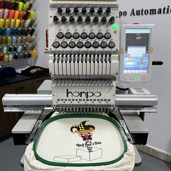 Eine Honpo-Stickmaschine stickt ein Design eines Narren auf einem Stoff. Die Maschine ist von verschiedenen Garnrollen umgeben.