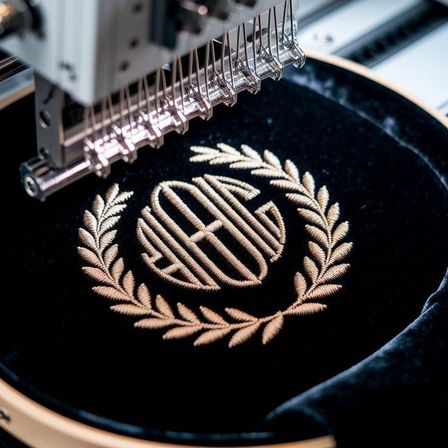 Eine Maschine stickt ein Monogramm auf einen schwarzen Samtstoff. Das Monogramm ist von einem Lorbeerkranz umgeben.