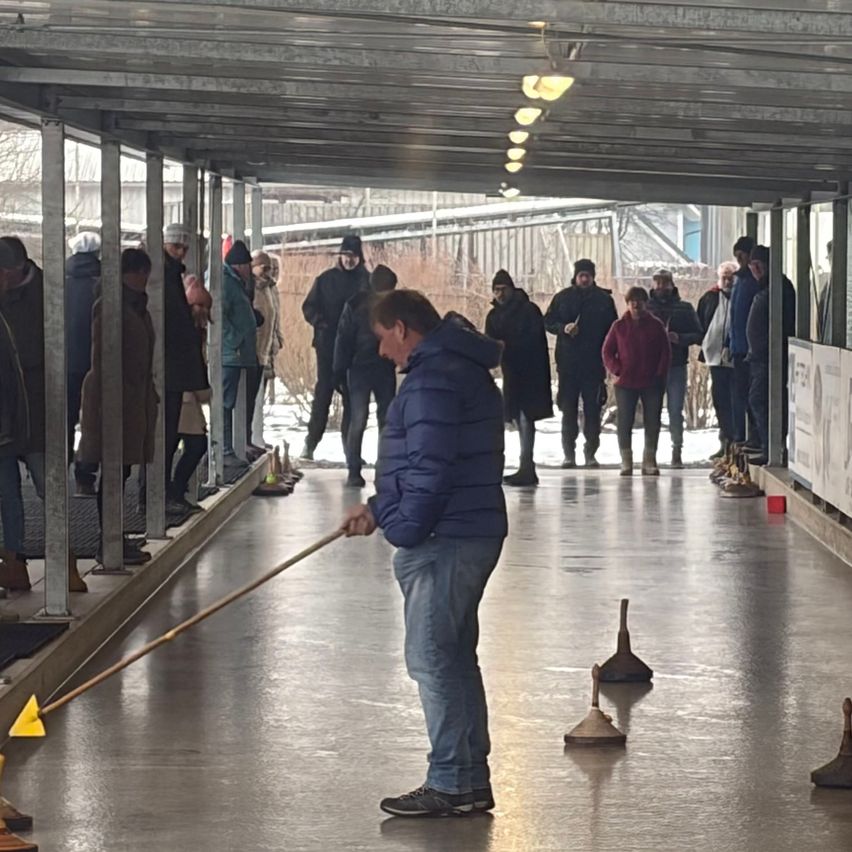 Ein Mann fegt den Boden einer überdachten Eisbahn. Mehrere Menschen stehen und warten darauf, zu spielen. Verkehrskegel und andere Gegenstände liegen auf dem Boden.
