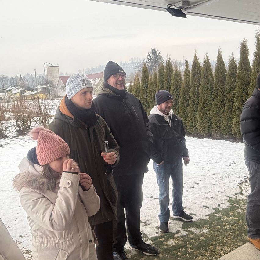 Eine Gruppe von Menschen, eingehüllt in Winterkleidung, steht in einer verschneiten Landschaft, wobei ein Mann eine Flasche hält und lächelt.