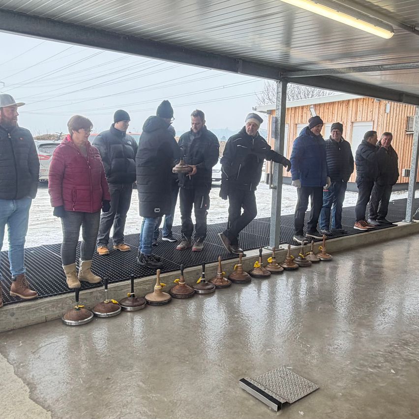 Eine Gruppe von Menschen in Winterkleidung steht in einem überdachten Bereich, einige halten Objekte. Der Boden ist nass und es sind mehrere Metallobjekte aufgereiht.