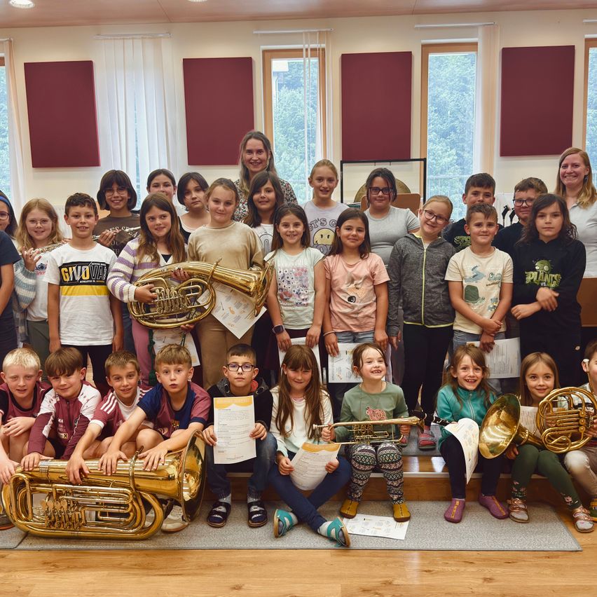 Eine Gruppe von Kindern, Lehrern und Erwachsenen posiert für ein Foto in einem Raum. Einige Kinder halten Urkunden und andere spielen Blechblasinstrumente. Der Raum hat einen Holzboden, einen grauen Teppich und Fenster.