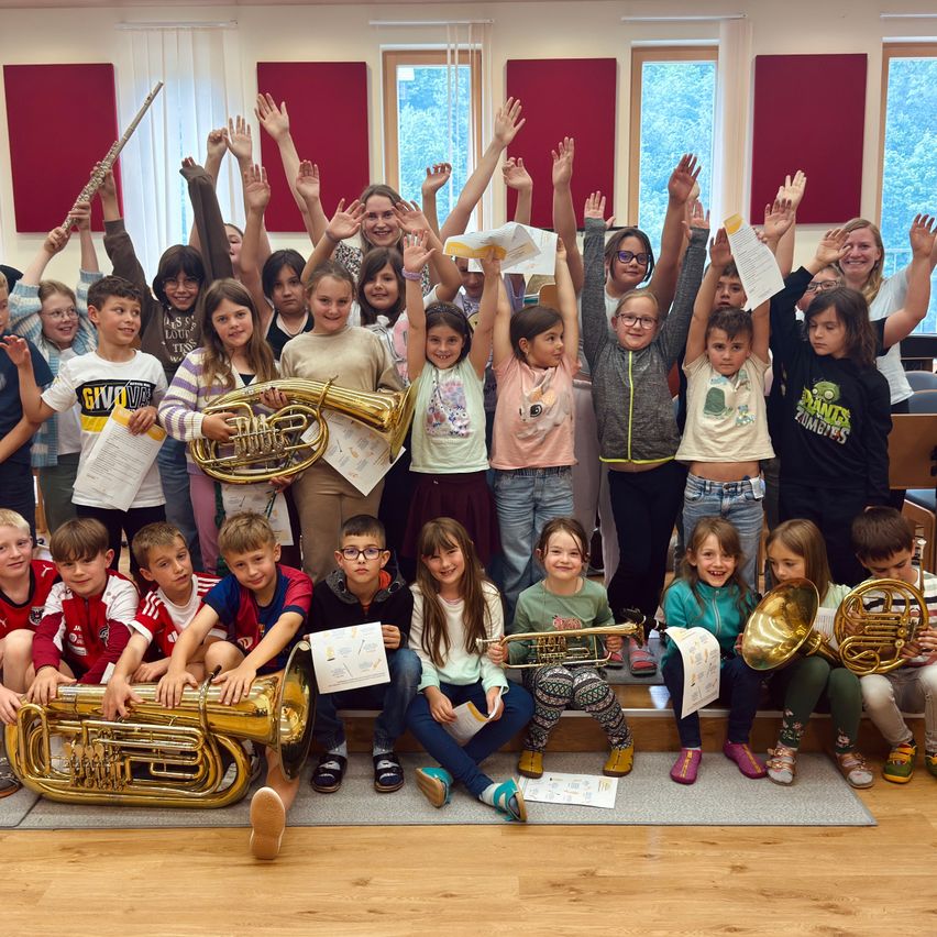Eine Gruppe von Kindern und Erwachsenen posiert mit Blechblasinstrumenten in einem Raum mit roten Wänden und großen Fenstern. Einige stehen und heben die Hände, andere sitzen auf dem Boden.