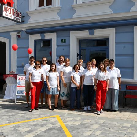 Eine Gruppe von Menschen in weißen Shirts und roten Hosen steht vor einem blauen Gebäude. Ein Banner mit der Nummer 200 ist links platziert.
