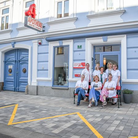 Eine Gruppe von Apothekern in weißen Kitteln und Brillen sitzt auf einer Bank vor einer Apotheke mit einem Schild, das 'Apotheke zum Hl. Leopold' steht.