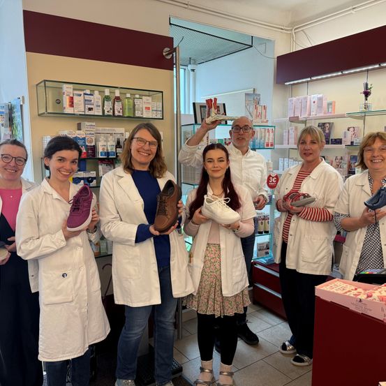 Eine Gruppe von Menschen in weißen Laborkitteln hält Schuhe in einer Apotheke. Sie scheinen zu lächeln und für ein Foto zu posieren.
