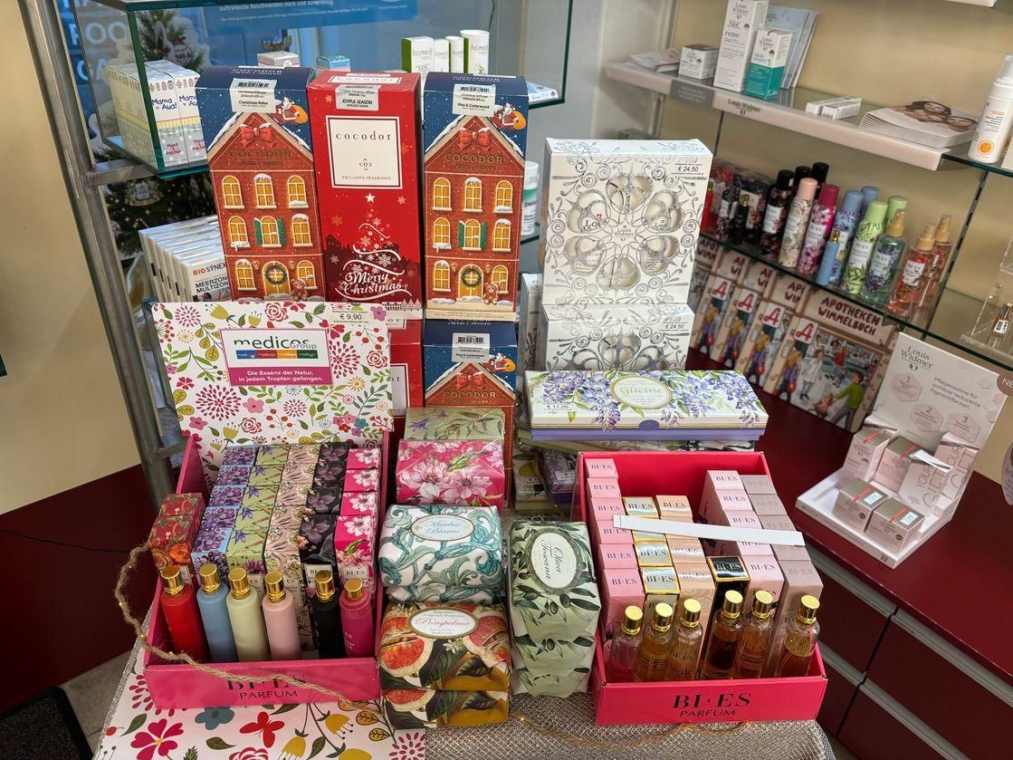 Eine Anzeige von Feiertags-Produkten in einem Geschäft, einschließlich Geschenkboxen, Kerzen und Flaschen. Die Produkte haben Blumen- und Weihnachtsdesigns. Im Hintergrund befinden sich auch Regale mit weiteren Produkten.