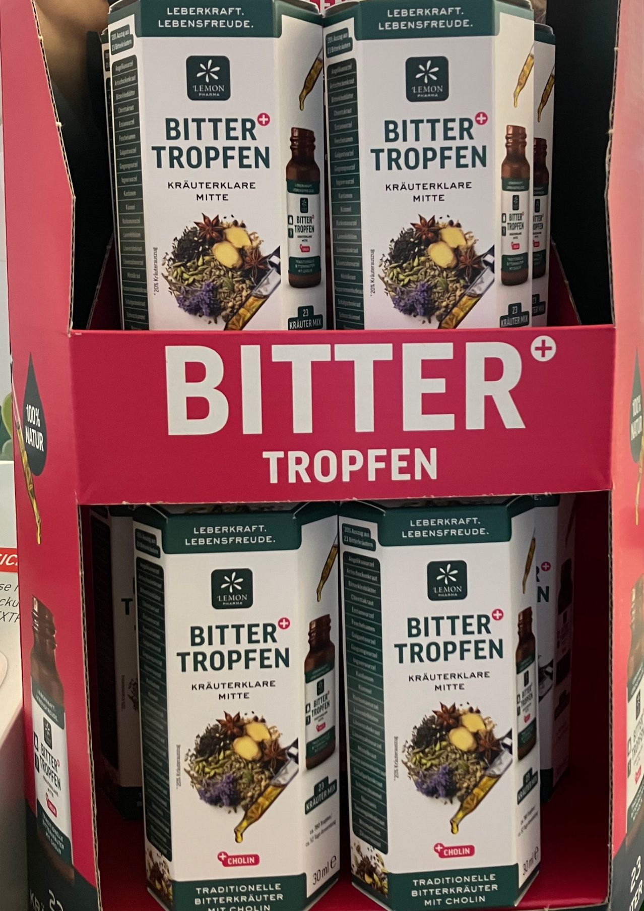 Ein roter Regal zeigt mehrere Schachteln von Bitter Tropfen, einem pflanzlichen Heilmittel, wobei das Wort 'BITTER' deutlich sichtbar ist. Die Schachteln sind grün und weiß und zeigen eine Illustration von Kräutern und Gewürzen.