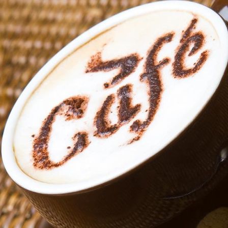 Eine Tasse Kaffee mit Milchschaumkunst oben, die 'Cafe' besagt. Die Tasse steht auf einem Untertasse mit einem dunkelbraunen Rand.