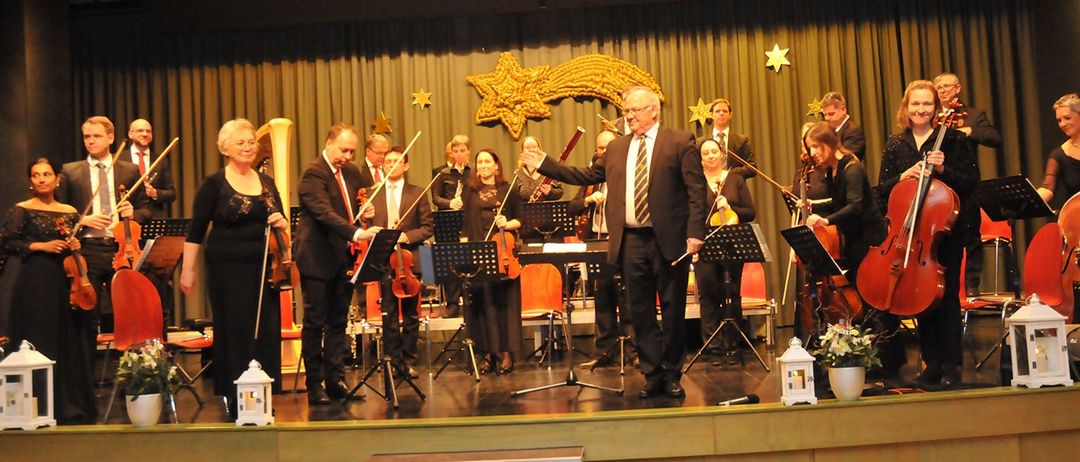 Eine Gruppe von Musikern spielt auf der Bühne mit Instrumenten wie Violinen, Celli und einer Harfe. Der Mann in der Mitte hält einen Dirigentenstab. Es gibt dekorative Sterne und ein sternförmiges Objekt auf der Bühne.