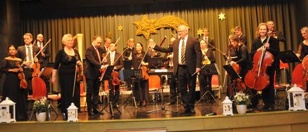 Eine Gruppe von Musikern spielt auf der Bühne mit Instrumenten wie Violinen, Celli und einer Harfe. Der Mann in der Mitte hält einen Dirigentenstab. Es gibt dekorative Sterne und ein sternförmiges Objekt auf der Bühne.