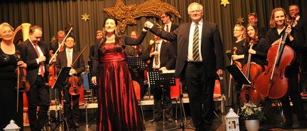 Eine Frau in einem roten Kleid und ein Mann in einem Anzug halten sich auf der Bühne die Hände, während ein Orchester im Hintergrund spielt.