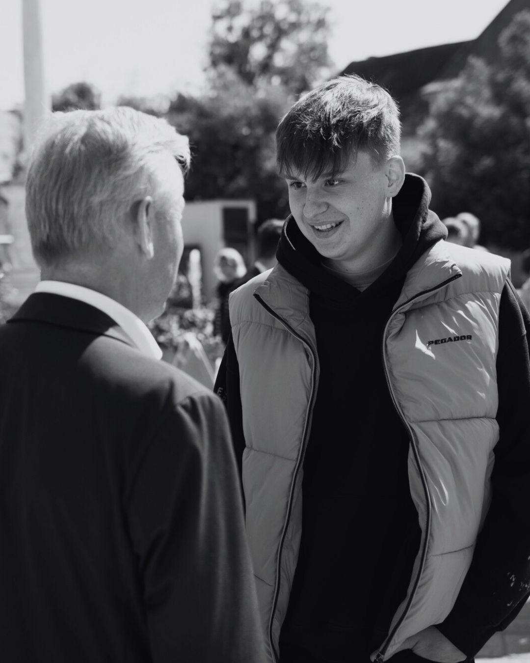 Ein Schwarz-Weiß-Foto zeigt einen Mann im Anzug, der mit einem lächelnden jungen Mann in einer gepolsterten Weste spricht.