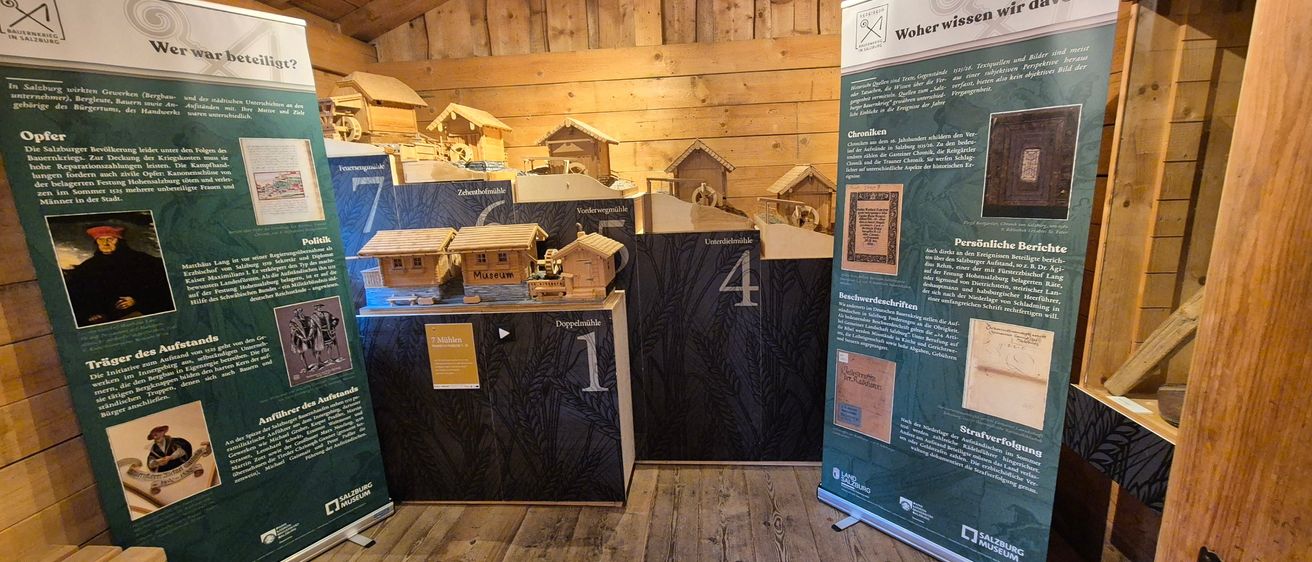 Eine hölzerne Ausstellung eines Dorfes mit verschiedenen Miniaturgebäuden. Es gibt nummerierte Schilder und Texttafeln, wobei ein Schild die Zahl eins zeigt. Holzwände sind im Hintergrund.