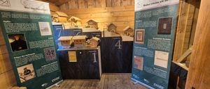 Eine hölzerne Ausstellung eines Dorfes mit verschiedenen Miniaturgebäuden. Es gibt nummerierte Schilder und Texttafeln, wobei ein Schild die Zahl eins zeigt. Holzwände sind im Hintergrund.