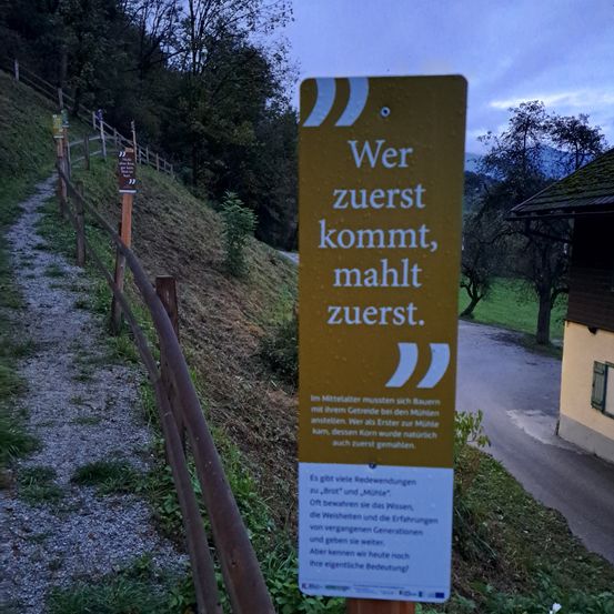 Ein gelbes Schild mit deutschem Text besagt 'Wer zuerst kommt, mahlt zuerst'. Darunter befindet sich ein kleiner Zitat. Der Hintergrund ist unscharf.