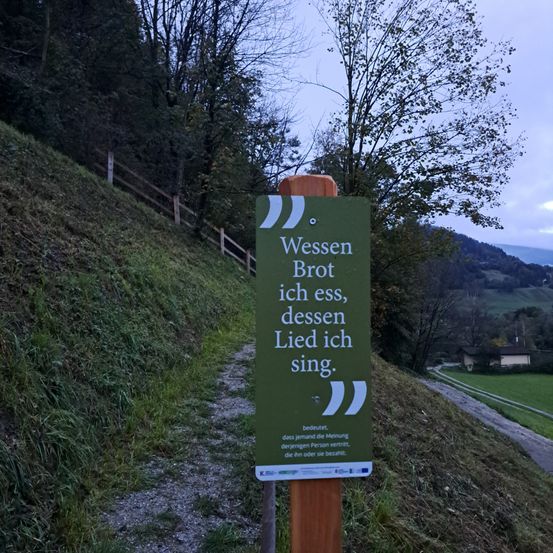Ein Schild auf einem Holzpfosten steht: 'Wenn ich esse, singe ich das Lied dessen, was ich esse.' Das Schild erklärt, dass die Meinung einer Person durch das, was sie isst oder trinkt, offenbart wird.