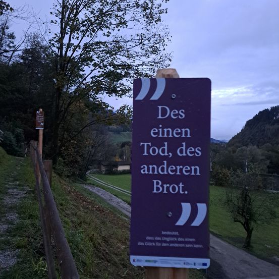 Ein lilafarbenes Schild mit einem Zitat darauf steht: 'Des einen Tod, des anderen Brot.' Das Schild befindet sich auf einem Grasweg in der Nähe eines Holzzauns.